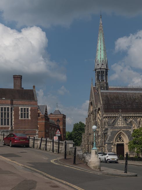 A view of a historic red-brick building with a prominent clock tower and ornate architectural features, situated behind a black wrought-iron gate with brick pillars topped by decorative lanterns, in a
